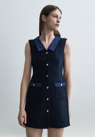 Femme portant une robe sans manches bleu marine avec un col bleu, des boutons sur le devant et deux poches avec rabats et boutons, debout devant un fond gris uni.