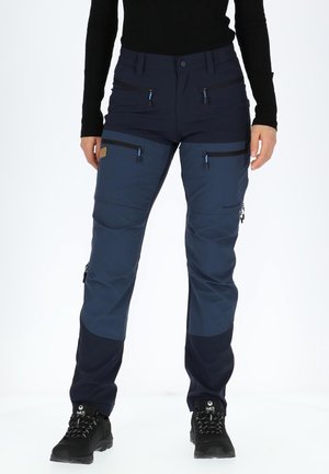 Frau trägt dunkelblaue Outdoor-Hose mit mehreren Reißverschlusstaschen und schwarzen Wanderschuhen, steht vor weißem Hintergrund.