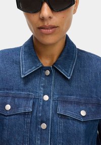 Chaqueta de mezclilla en azul oscuro con cuello, botones a presión plateados y bolsillos en el pecho. La tela parece texturizada y duradera.