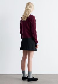 Bordeauxfarbenes Sweatshirt, schwarzer Rock, graue Knöchelsocken und schwarze Slipper. Das Modell wird von hinten betrachtet und steht vor einem einfarbigen hellen Hintergrund.