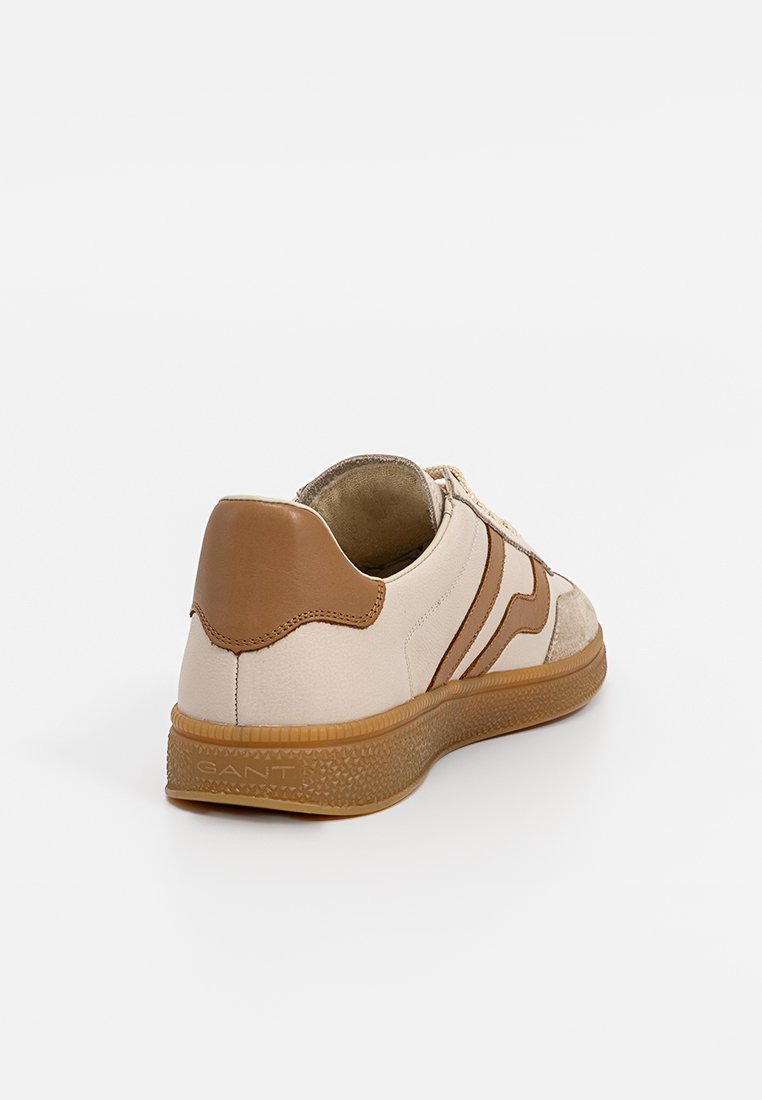 Sneaker beige in pelle con accenti marroni, dotato di una suola testurizzata e un tallone sollevato. Il lato presenta un motivo a strisce ondulate.