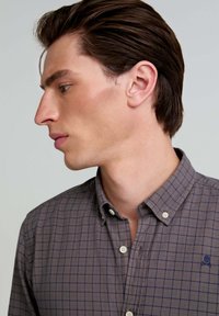 Camisa de cuadros en marrón y gris con cuello, botones blancos y un pequeño logotipo bordado en el pecho. Textura suave.