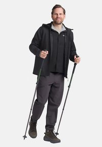 Veste noire sur une chemise grise, pantalon gris et bottes marron. L'utilisateur tient des bâtons de randonnée, debout avec une expression neutre.