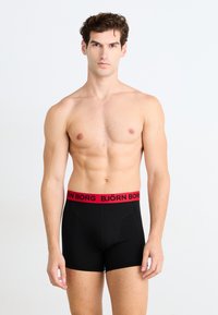 Boxers noirs avec une taille rouge portant le texte "BJÖRN BORG". Tissu lisse, design ajusté, longueur mi-cuisse.