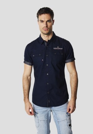 Camisa azul marino de manga corta con botones, dos bolsillos en el pecho, mangas remangadas y detalle de logo en el hombro; textura de tela suave.