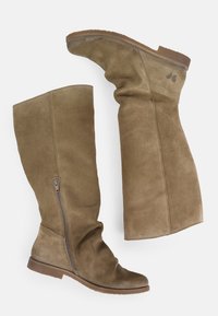 Bottes en daim marron jusqu'au genou avec fermeture éclair latérale et semelles en caoutchouc plates, une botte debout et l'autre couché sur le côté.