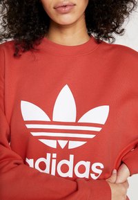 Röd sweatshirt med vitt Adidas-logotyp, med en stor trefoil och tre horisontella ränder. Mjuk tyg med ribbat krage och muddar.