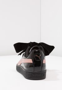 Černé lesklé tenisky Puma s růžovým bočním proužkem, velkou černou sametovou mašlí na tkaničkách a texturovanou černou podrážkou na bílé ploše.