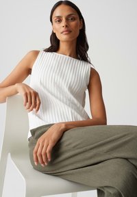 Top in cotone bianco a righe con scollatura quadrata, abbinato a pantaloni larghi verde oliva dalla trama texture. Il modello è seduto su una sedia bianca.