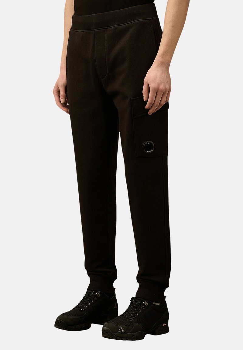 Schwarze Jogginghosen aus weichem Stoff, mit einer Seitentasche, elastischem Bund und sichtbaren Nähdetails. Kombiniert mit schwarzen Schuhen.