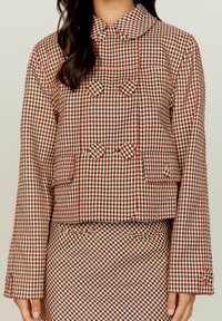 Marynarka w kratę z kołnierzykiem, cechująca się połączeniem czerwonych, brązowych i białych kwadratów, podwójnym zapięciem i kieszeniami z przodu.