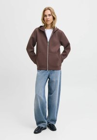 Felpa marrone con zip e tasche frontali, abbinata a jeans a gamba larga blu chiaro e scarpe nere, su uno sfondo neutro.