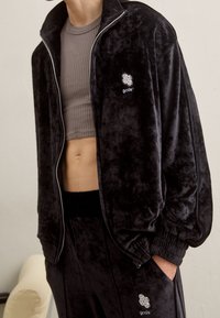 Zwarte fluwelen trainingspak met een jas met ritssluiting en een geribd cropped topje. Voorzien van elastische manchetten en tailleband, met een logodetail op de borst.