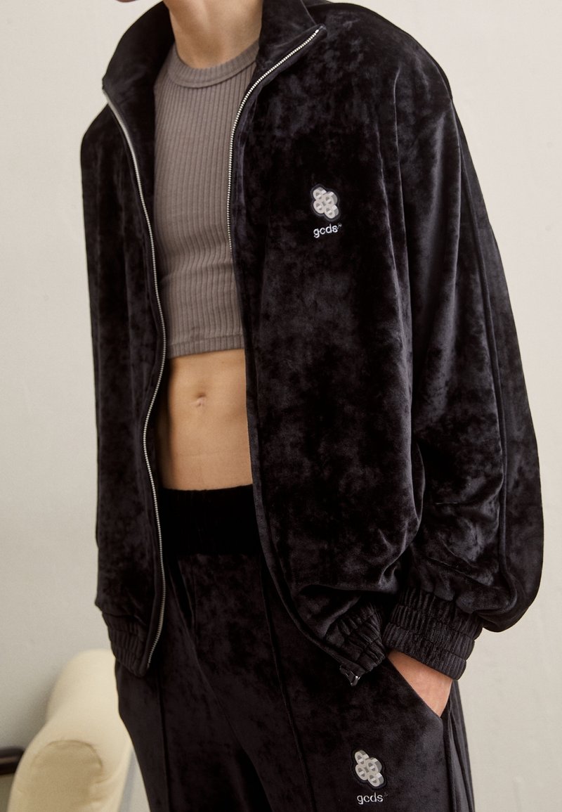 Zwarte fluwelen trainingspak met een jas met ritssluiting en een geribd cropped topje. Voorzien van elastische manchetten en tailleband, met een logodetail op de borst.