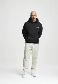 Mustelukollane kapuutsiga särk, millel on ees tasku ja valge tekst, koos kreemja dressipüksiga ja valgete tossudega. Sile kangas, lahtine lõige.