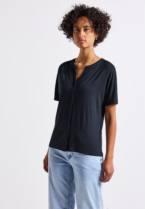Schwarzes kurzärmliges Hemd mit Knöpfen aus glattem Stoff, mit V-Ausschnitt und lässiger Passform. Ein Paar hellblaue Jeans ist sichtbar.