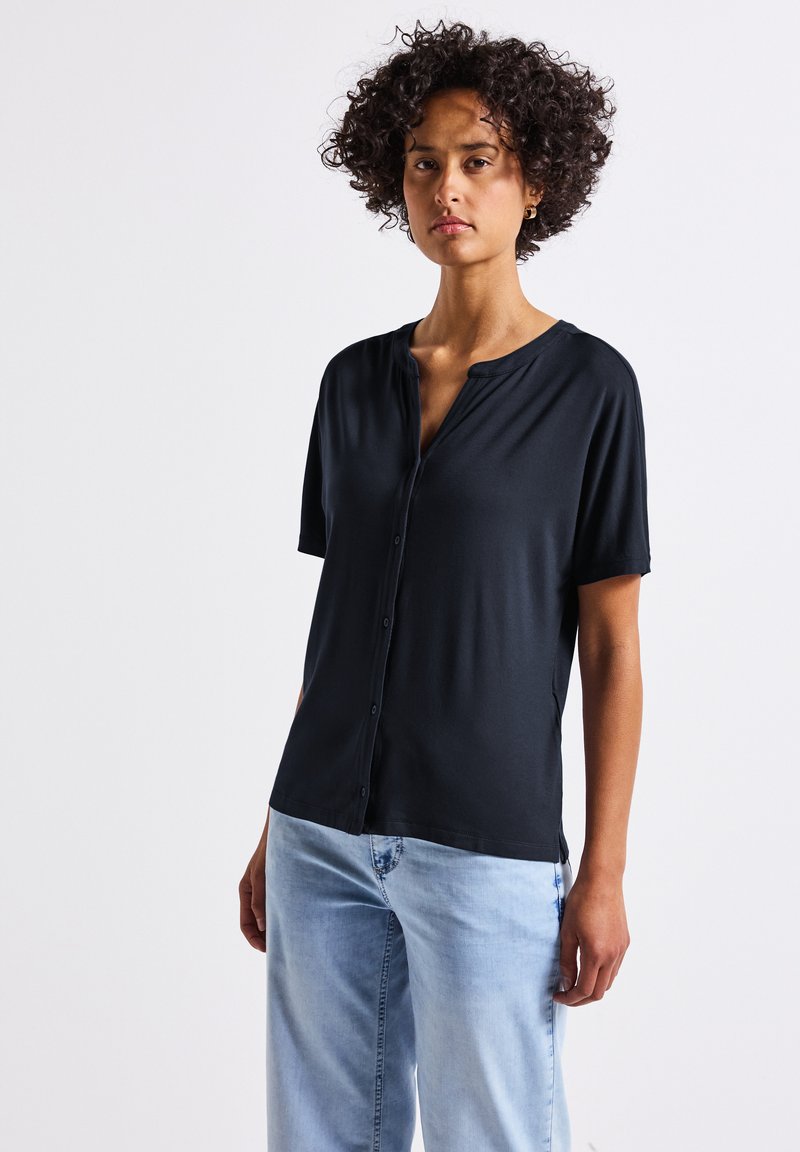 Camicetta nera a maniche corte con bottoni realizzata in tessuto morbido, caratterizzata da un collo a V e una vestibilità comoda. Sono visibili dei pantaloni in denim blu chiaro.