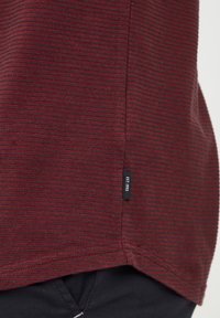 Burgundy striped textile with a short side slit. Features a label on the hem that reads "Established 1993." Fabric has a textured finish.