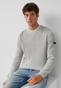 Hellgrauer, gerippter Strickpullover mit Rundhalsausschnitt, strukturiertem Stoff und einem schwarzen Logopatch an der linken Ärmels. Kombiniert mit blauen Jeans.