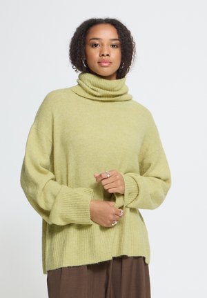Femme aux cheveux bouclés portant un pull à col roulé ample vert clair et un pantalon marron, tenant une manche avec ses mains.