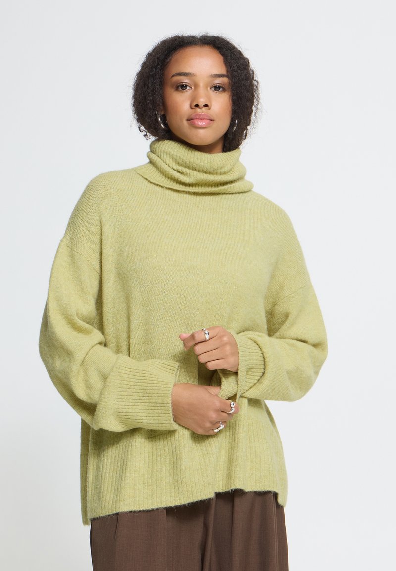 Femme aux cheveux bouclés portant un pull à col roulé ample vert clair et un pantalon marron, tenant une manche avec ses mains.