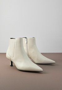 Bottines blanches en cuir avec bouts pointus, fermeture éclair sur le côté, accents en élastique nervuré et un petit talon en kitten noir sur un fond neutre.