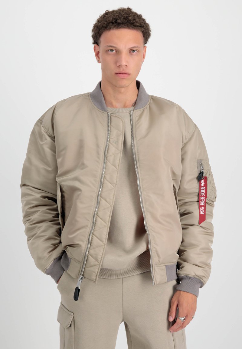 Chaqueta bomber beige con cremallera frontal, puños acanalados y una etiqueta roja en la manga. Tela texturizada y detalles de diseño acolchados.