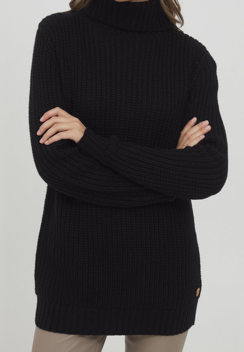 Personne portant un pull à col roulé noir en grosse maille avec manches longues, les bras croisés, et un pantalon beige sur un fond neutre.