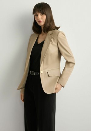 Kobieta o brązowych włosach, ubrana w beżową marynarkę na czarnym topie i czarnych spodniach, stojąca na tle gładkiej jasnej ściany.