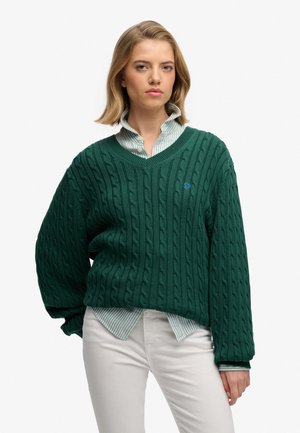 Pull en tricot vert avec un motif en relief, design en col en V et un petit logo bleu. Porté par-dessus une chemise rayée bleu clair et un pantalon blanc.
