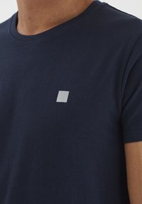 Tumši zilas krāsas T-krekls ar apaļu kakla izgriezumu, izgatavots no kokvilnas. Uz kreisā krūtīm ir neliels balts svītrots kvadrātveida logo.