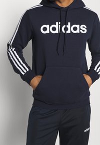 Hoodie bleu marine avec le logo "adidas" blanc, des cordons de serrage et une poche avant ; présente trois bandes blanches sur les manches. Matière douce en mélange de coton.