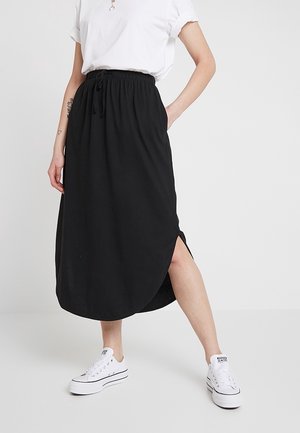 Femme portant une jupe midi noire fendue sur le côté, des baskets blanches et un haut blanc à manches courtes, debout avec une main dans la poche.