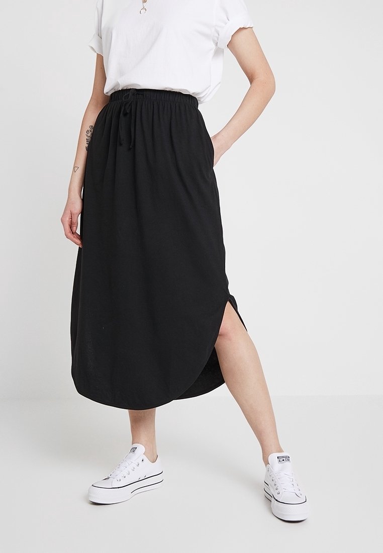 Femme portant une jupe midi noire fendue sur le côté, des baskets blanches et un haut blanc à manches courtes, debout avec une main dans la poche.