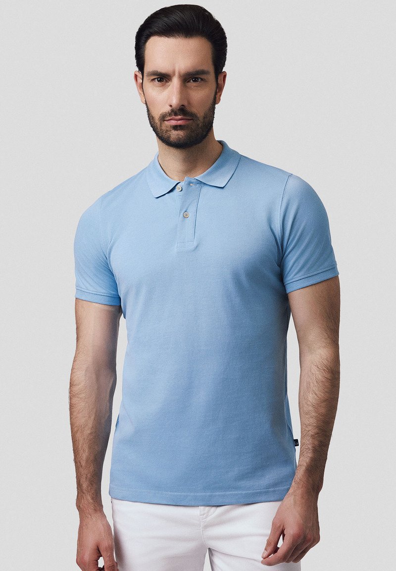 Jasnoniebieska koszulka polo wykonana z bawełny, z klasycznym kołnierzykiem, krótkimi rękawami i zapinana na trzy guziki. Widoczne są również białe spodnie.