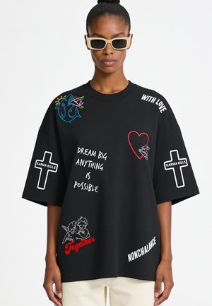 Frau trägt ein schwarzes übergroßes T-Shirt mit weißem und rotem Text und Grafiken, darunter die Kreuze "Dream Big Anything Is Possible" und "Karma Kills".