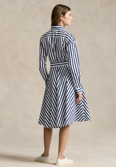 Nő, aki kék-fehér függőleges csíkos, hosszú ujjú midi ruhát visel, amelyhez illő csíkos öv és fehér sportcipő tartozik, oldalra állva.