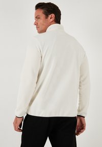 Hvid fleece-pullover med høj krave, der har en glat tekstur og sort kant ved kanten og ærmerne. Vist fra bagsiden.