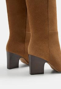 Bottines en daim marron avec une finition lisse, bout carré, et talon bloc en bois de hauteur moyenne. Sans ornements ni motifs visibles.