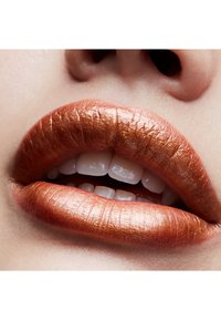 Rouge à lèvres en cuivre métallique sur des lèvres bien dessinées, mettant en valeur une texture lisse et un éclat. Dents blanches visibles, teint de peau naturel en arrière-plan.