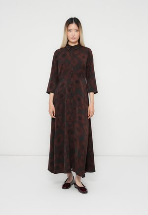 Woman standing against white background wearing a long dark dress with red floral patterns and burgundy ballet flats.