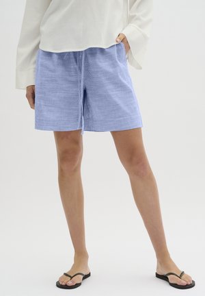 Kobieta w jasnoniebieskich, teksturowanych szortach, białej bluzce z długim rękawem i czarnych japonkach, stojąca z jedną ręką w kieszeni na białym tle.