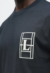 Κοντινό πλάνο σκούρου ναυτικού πουκάμισου με λευκό γεωμετρικό λογότυπο που περιλαμβάνει ένα μεγάλο γράμμα «L» στο στήθος.