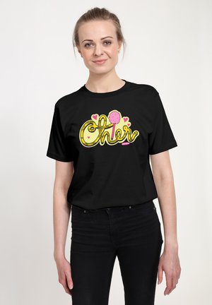Kobieta ubrana w czarną koszulkę z żółtym napisem "Cheer", różowym pomponem i serduszkami, w połączeniu z czarnymi spodniami, stoi na białym tle.