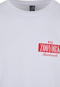 White cotton t-shirt with a round neckline featuring red printed text: "N.Y.C. ZOO YORK Skateboards." Smooth texture, standard fit.