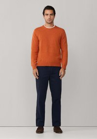 Pomarańczowy sweter z dzianiny z okrągłym dekoltem, noszony na granatowych spodniach. Ciemnobrązowe buty dopełniają strój. Prosty design, bez wzorów ani akcentów.