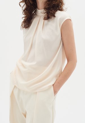 Vrouw draagt een mouwloze crèmekleurige blouse met geplooide halslijn en bijpassende crèmekleurige broek met handen in de zakken.
