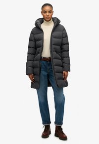 Mørkegrå pufferjakke med hætte, der har en quiltet design og sidelommer, parret med en lysfarvet turtleneck og blå jeans.