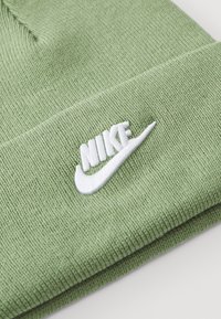 Zelena pletena kapa z mehkimi vlakni, z belim vezenim logotipom Nike in zasnovo z zavihkom.