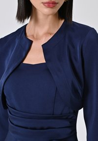 Giacca cropped in satin blu navy con scollo arrotondato e design aderente, abbinata a un vestito aderente coordinato. Texture e cuciture lisce.